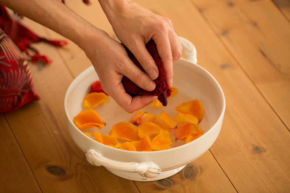 Eine Frauenhand drückt einen Waschlappen über einer Schüssel mit Wasser und Rosenblütenblättern aus.